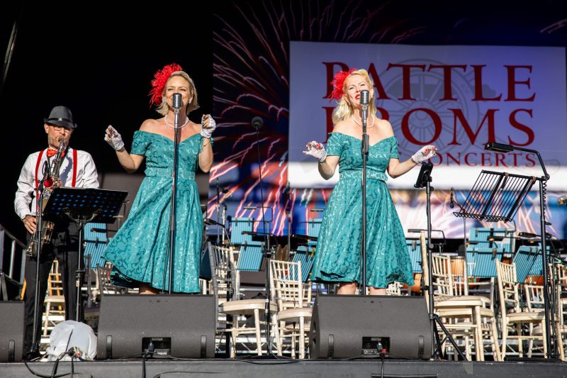 Performers singing at Battle Proms concert with stage backdrop.