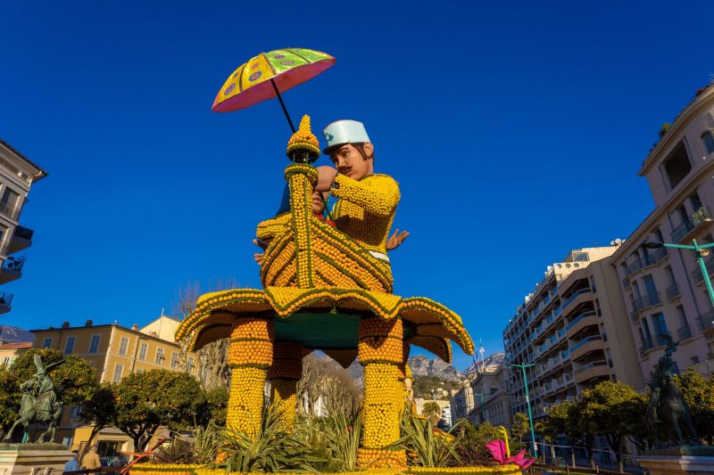 Colorful parade float with a large figure holding an umbrella in a city square.