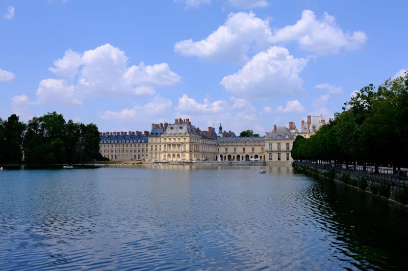 View of a historic French palace with a large water body in front and a blue sky with clouds.