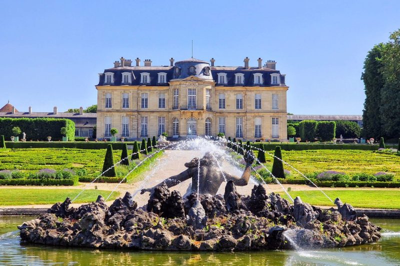 French château with fountain and manicured gardens in bright sunlight.